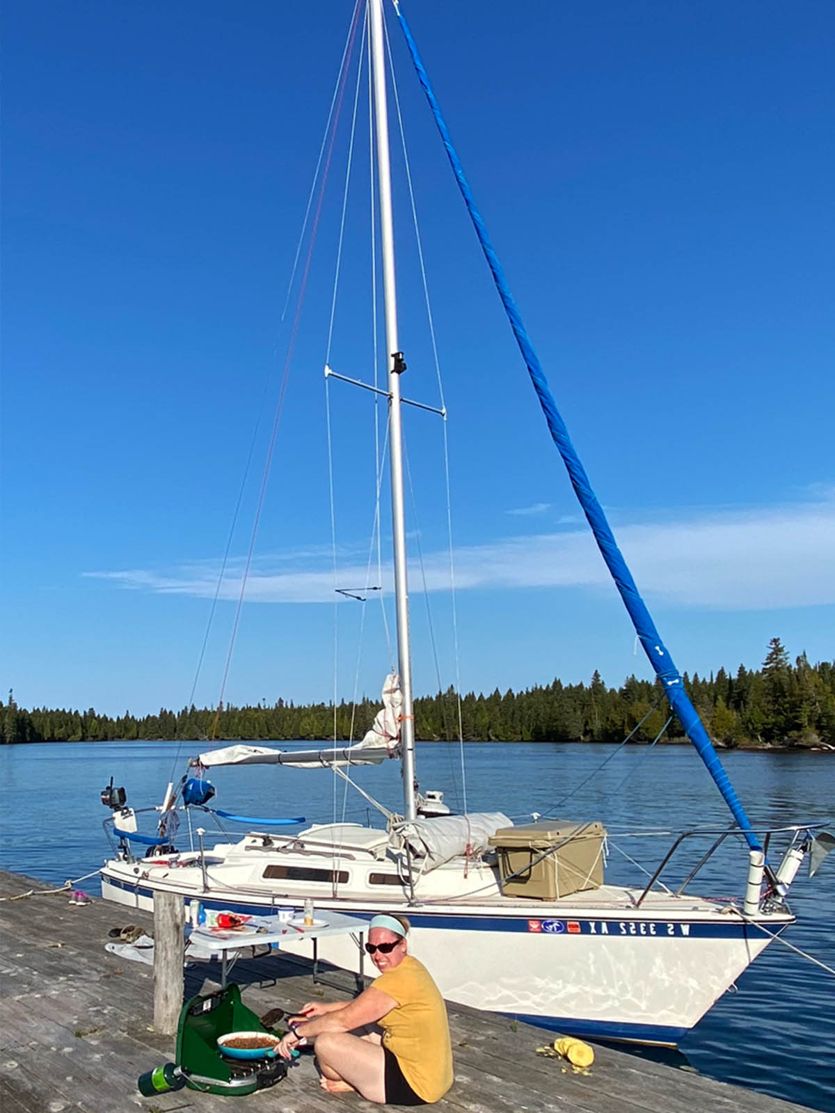 sarah sitting on dock in front of sailboat cooking on campstove